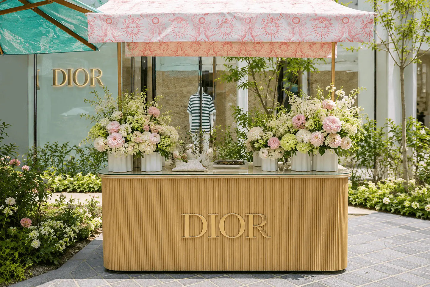 A Dior-branded floral display stand in an outdoor garden setting, featuring a pleated gold-toned base, a pink patterned canopy, and multiple arrangements of pink, peach, and white flowers.