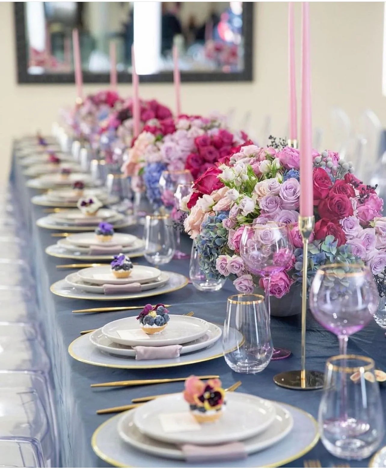 A long banquet table set for guests with layered white plates, gold-rimmed chargers, pink and purple floral centerpieces, tall pink taper candles, and clear wine glasses on a blue tablecloth.