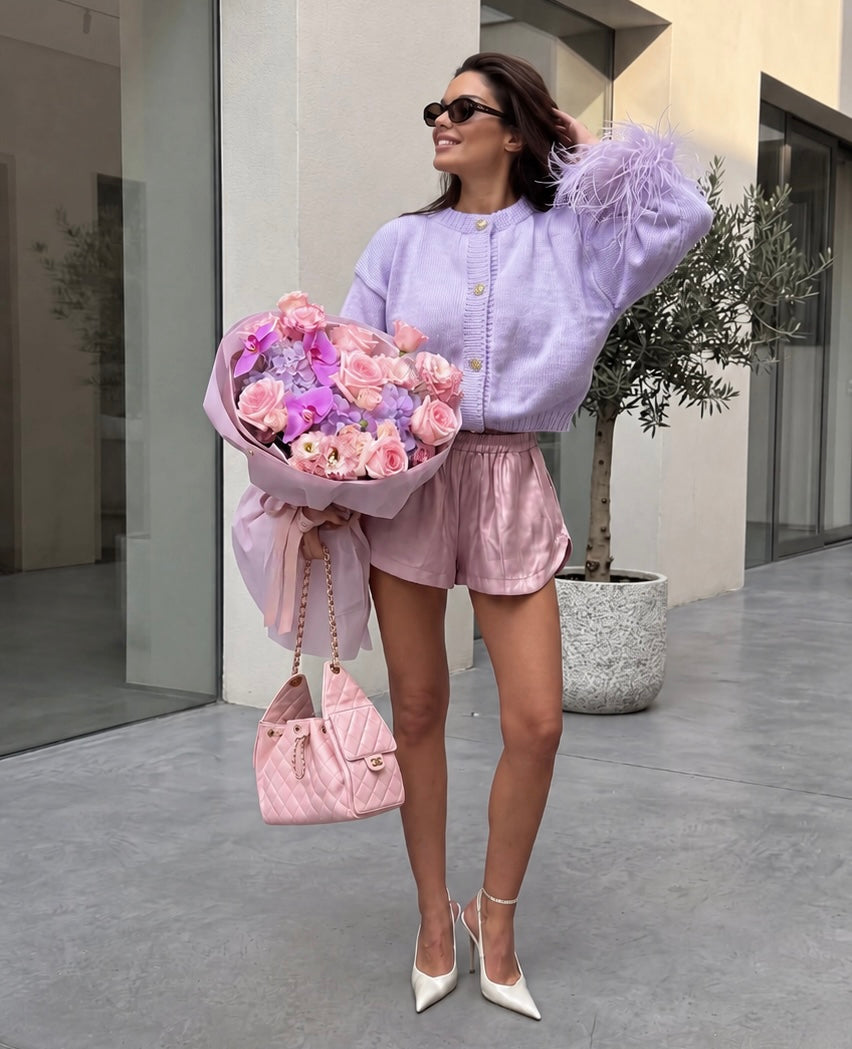 A woman stands outdoors holding a large bouquet of pink and purple flowers, wearing a light lavender cardigan with feathered sleeves, shiny pink shorts, white pointed heels, and carrying a pink quilted handbag.